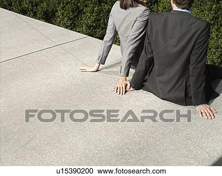 Stock Image - Business man and woman sitting on wall outdoors back view (cropped). Fotosearch