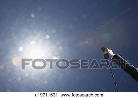 Stock Image - Business man holding telephone receiver against sky close up of hand. Fotosearch