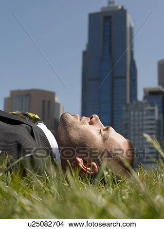 Business man lying on grass office buildings in background View Large Photo Image Picture - Business man lying on grass office buildings in background. Fotosearch