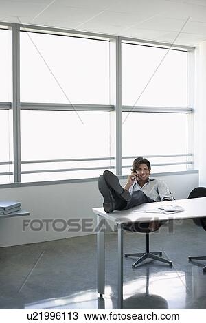 Stock Image - Business man using mobile phone sitting at office desk with feet up. Fotosearch