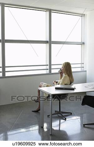 Stock Photography - Business woman drinking from cup sitting in office. Fotosearch