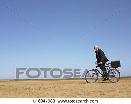 Stock Image - Businessman riding bicycle in desert. Fotosearch