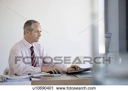 Businessman sitting at desk in office using computer. View Large Photo Image Stock Image - Businessman sitting at desk in office using computer.. Fotosearch