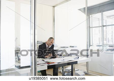 Stock Image - Businessman sitting at desk in office talking on phone.. Fotosearch