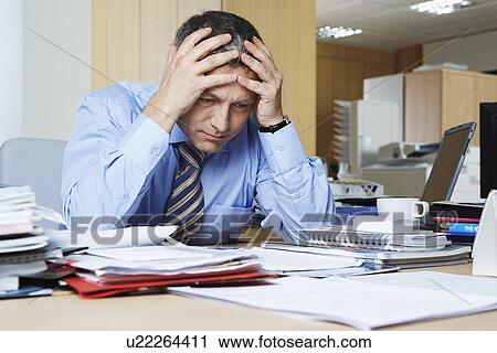 Stock Image - Businessman sitting at desk with head in hands. Fotosearch