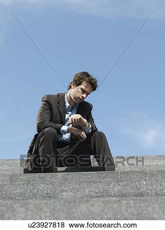 Stock Photo - Businessman sitting on steps outdoors looking at wristwatch low angle view. Fotosearch