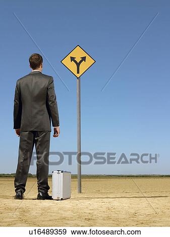 Businessman standing by briefcase looking at road sign in desert back view View Large Photo Image Stock Photo - Businessman standing by briefcase looking at road sign in desert back view. Fotosearch