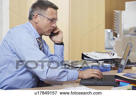 Stock Photo - Businessman talking on mobile phone using laptop in office. Fotosearch