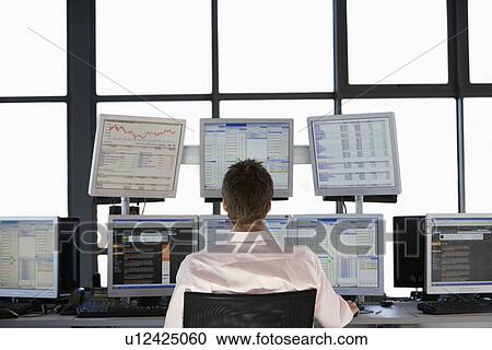 Stock Image - Businessmen sitting at desk watching computer screens back view.. Fotosearch