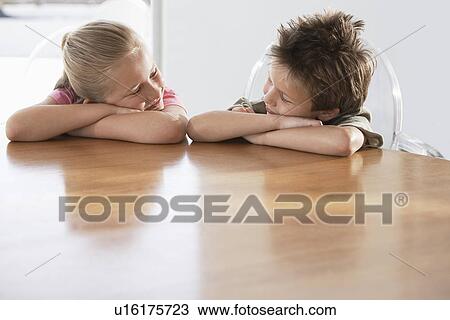 Stock Image - Children sitting in chairs leaning heads on table. Fotosearch