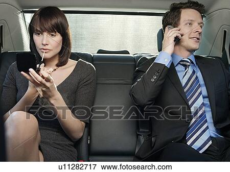 Stock Photo - Couple at back seat of car woman applying make-up man using mobile phone. Fotosearch