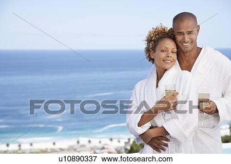 Stock Photo - Couple in bathrobes holding drinks embracing ocean in background. Fotosearch