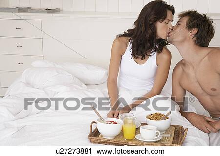 Stock Image - Couple kissing and having breakfast in bed. Fotosearch