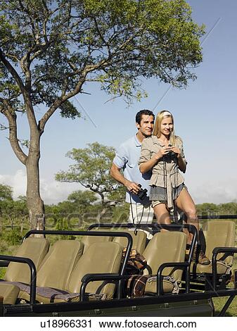 Stock Image - Couple on safari standing in jeep woman holding binoculars. Fotosearch