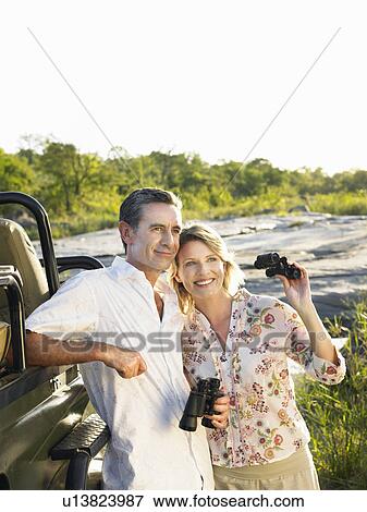 Stock Photo - Couple standing by jeep holding binoculars smiling. Fotosearch