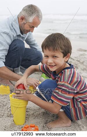 Stock Image - Father and son (5-6) playing in sand on beach. Fotosearch