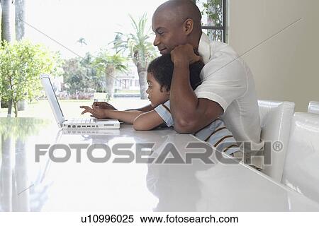 Stock Photography - Father and son (5-6 years) using laptop in dining room side view. Fotosearch