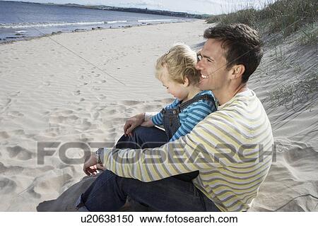 Stock Image - Father embracing daughter (5-6) sitting on beach. Fotosearch