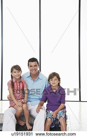 Stock Image - Father with son (7-9) and daughter (5-6) sitting indoors. Fotosearch