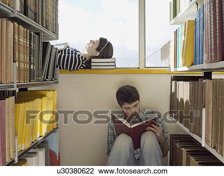 Stock Image - Female student lying down on library windowsill male student sitting on library floor reading. Fotosearch