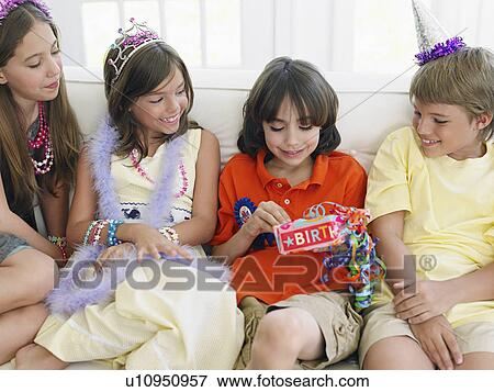 Stock Photo - Four children (7-12) sitting on sofa at birthday party one opening present. Fotosearch
