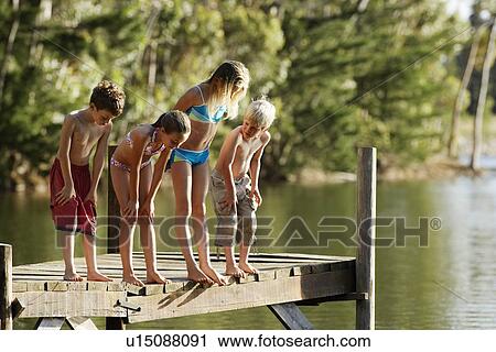 Stock Image - Four children (7-9) in swimwear standing on dock by lake.. Fotosearch