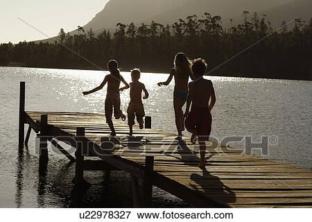 Stock Photo - Four children (7-9) running down dock by lake back view.. Fotosearch
