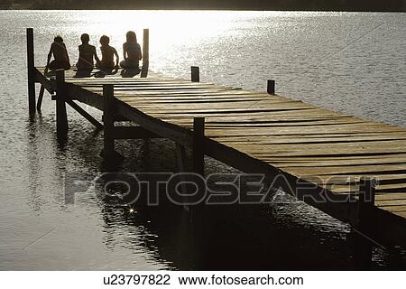 Stock Image - Four children (7-9) sitting on dock by lake back view.. Fotosearch