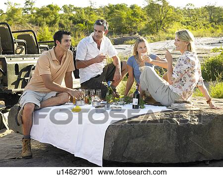 Stock Image - Four people having picnic on rock trees in background. Fotosearch