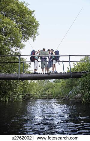 Stock Image - Four teenagers (16-17 years) backpacking in forest reading map on bridge. Fotosearch