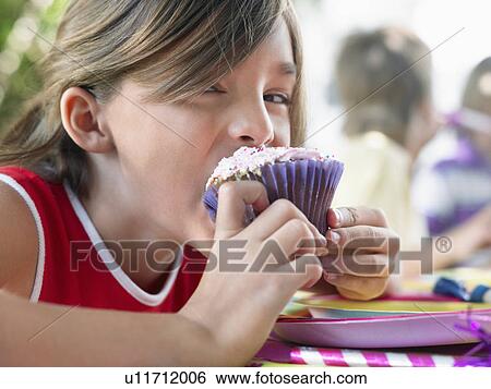Stock Photograph - Girl (10-12) eating cupcake at birthday party. Fotosearch