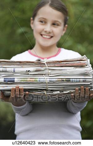 Stock Image - Girl (10-12) holding bundle of waste paper smiling looking away (focus on pile). Fotosearch
