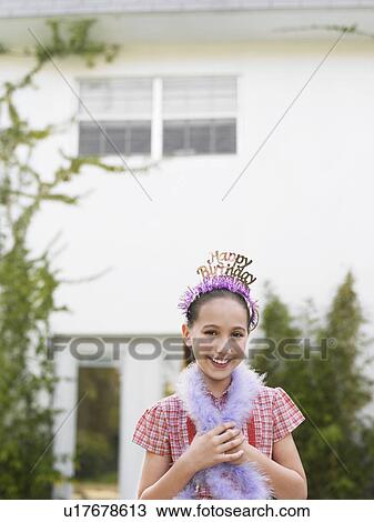Stock Image - Girl (10-12) in tiara and feather boa standing outside house. Fotosearch