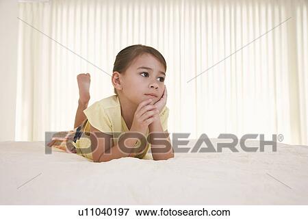 Stock Photo - Girl (5-6) lying on bed looking away. Fotosearch