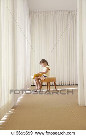 Stock Photo - Girl (5-6) sitting on stool in room reading book side view. Fotosearch