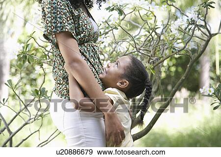 Stock Photo - Girl (5-6 years) hugging mother standing in garden side view. Fotosearch