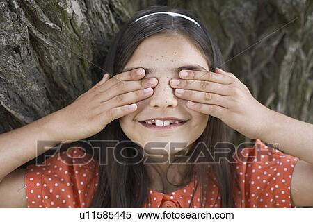 Stock Photography - Girl (7-9) playing hide and seek covering eyes by tree. Fotosearch