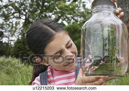 Stock Image - Girl (7-9) with eyes closed holding jar of stick insects close-up. Fotosearch