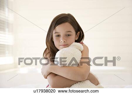 Stock Image - Girl cuddling teddy bear sitting on bed half length. Fotosearch