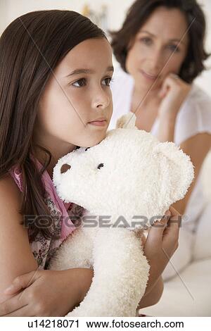 Stock Image - Girl hugging teddy bear half length mother in background. Fotosearch