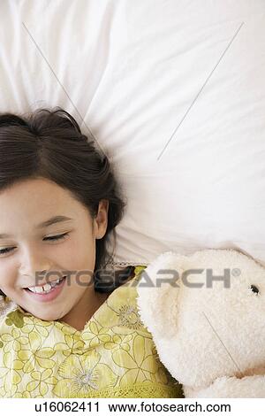 Stock Image - Girl lying on bed with teddy bear smiling head and shoulders. Fotosearch