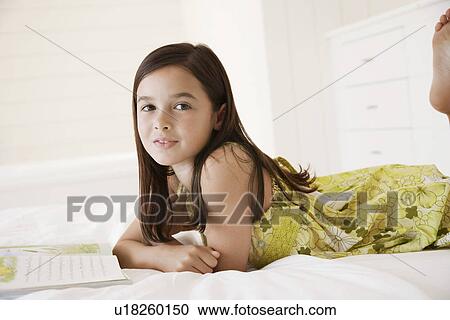 Stock Image - Girl reading story book lying on stomach on bed. Fotosearch
