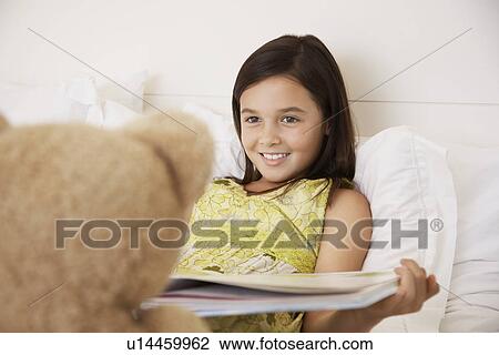Stock Image - Girl reading story to teddy bear sitting on bed. Fotosearch