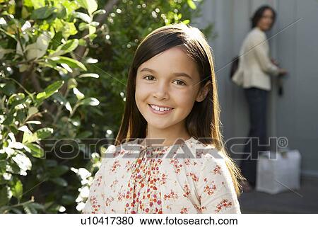 Stock Image - Girl smiling head and shoulders mother locking door in background. Fotosearch