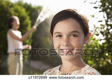 Stock Photo - Girl smiling head and shoulders mother watering garden. Fotosearch