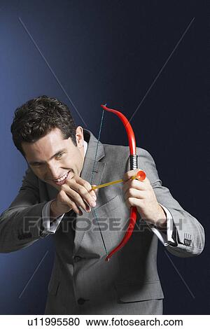 Stock Image - Man aiming toy bow and arrow against dark background. Fotosearch