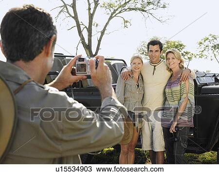Stock Image - Man and two women posing by jeep man taking photograph. Fotosearch