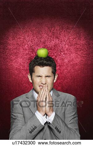 Stock Image - Man cringing with apple on head against red velvet background. Fotosearch
