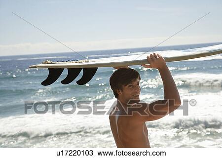 Stock Image - Man holding surfboard above head by ocean head and shoulders. Fotosearch
