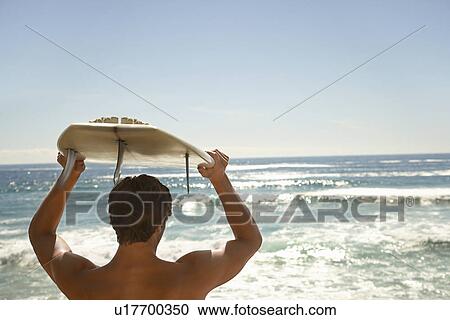 Stock Image - Man holding surfboard above head by ocean back view head and shoulders. Fotosearch